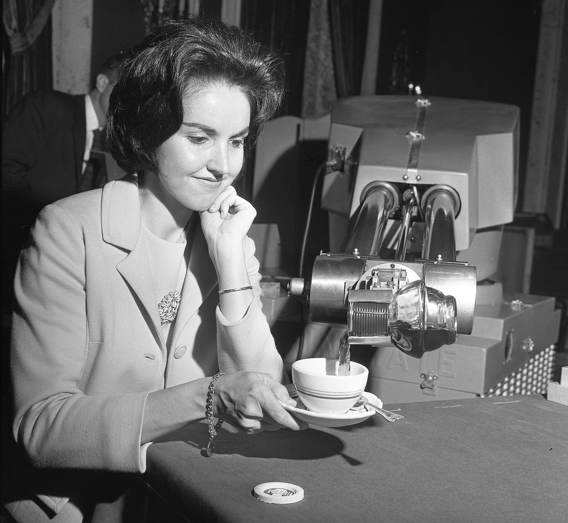 A Unimate industrial robot arm pours a cup of coffee for a seated woman at a 1967 hotel demonstration.